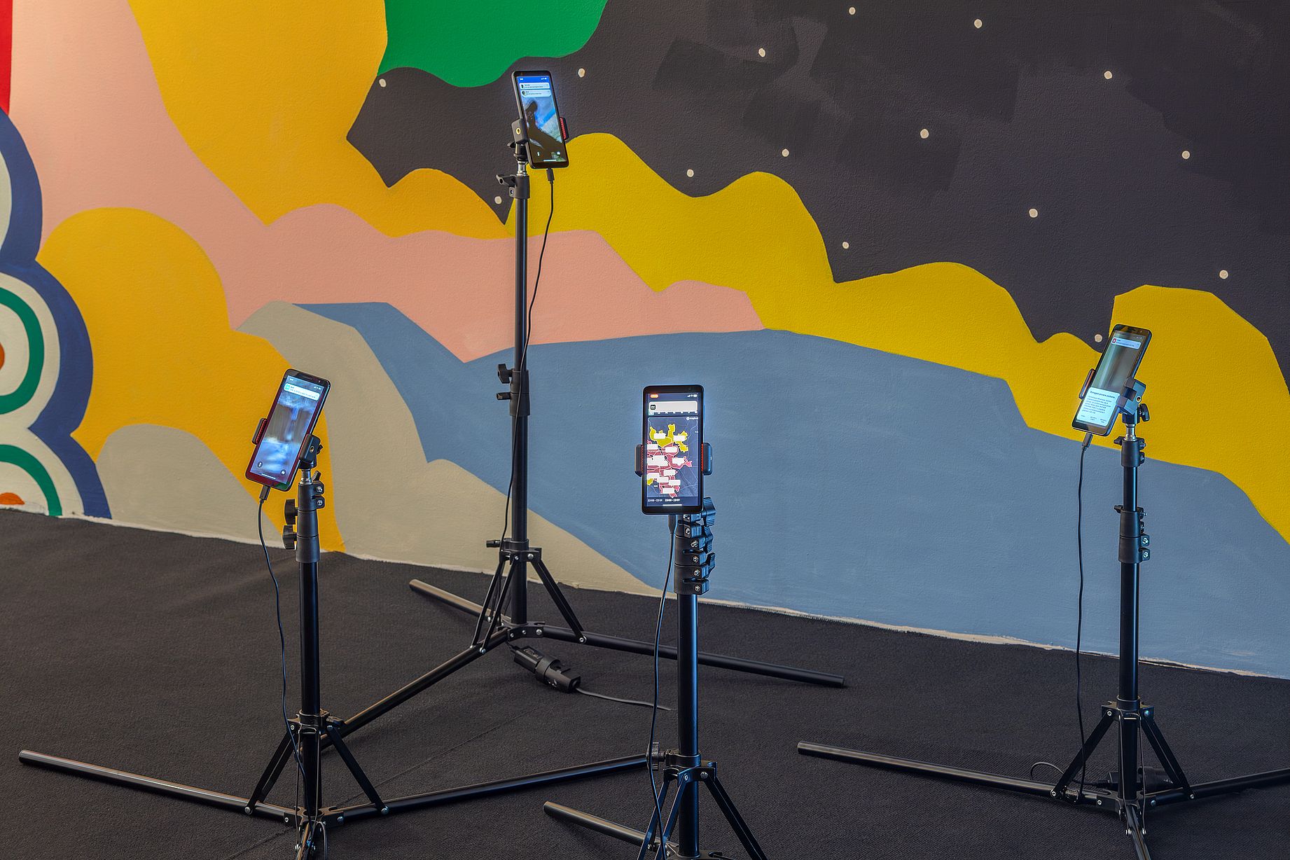 Wielobarwny mural autorstwa kolektywu Aravani Art Project przedstawia kilka kobiet w tradycyjnych strojach, otoczonych dużymi kwiatami i ptakiem, w pomieszczeniu zaaranżowanym z poduszkami i patchworkowymi dywanami na podłodze. 