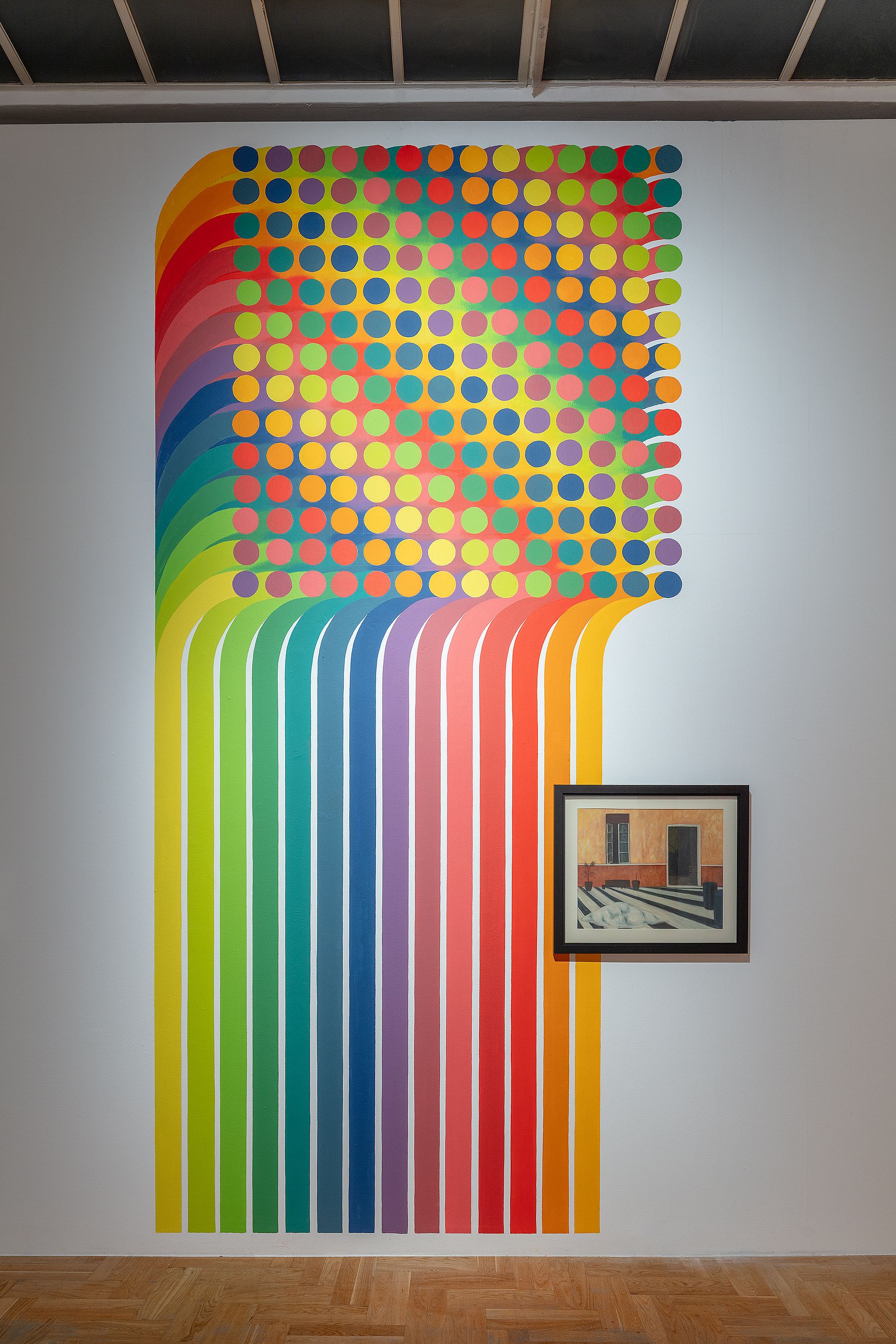Na białej ścianie galerii sztuki znajduje się duża, abstrakcyjna instalacja ścienna w kolorach tęczy, składająca się z pionowych pasów przechodzących w siatkę wielobarwnych kropek, a poniżej wisi mały, oprawiony obraz przedstawiający fasadę budynku.