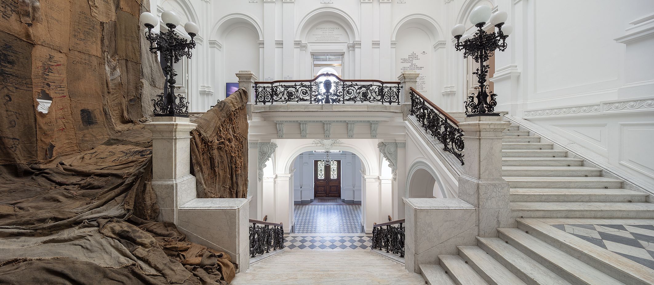 Obraz przedstawia elegancką, marmurową klatkę schodową wewnątrz Galerii Narodowej Zachęta w Warszawie, ozdobioną kutymi żelaznymi balustradami, białymi, arkadowymi ścianami i dużą, opadającą brązową instalacją artystyczną z tkaniny po lewej stronie