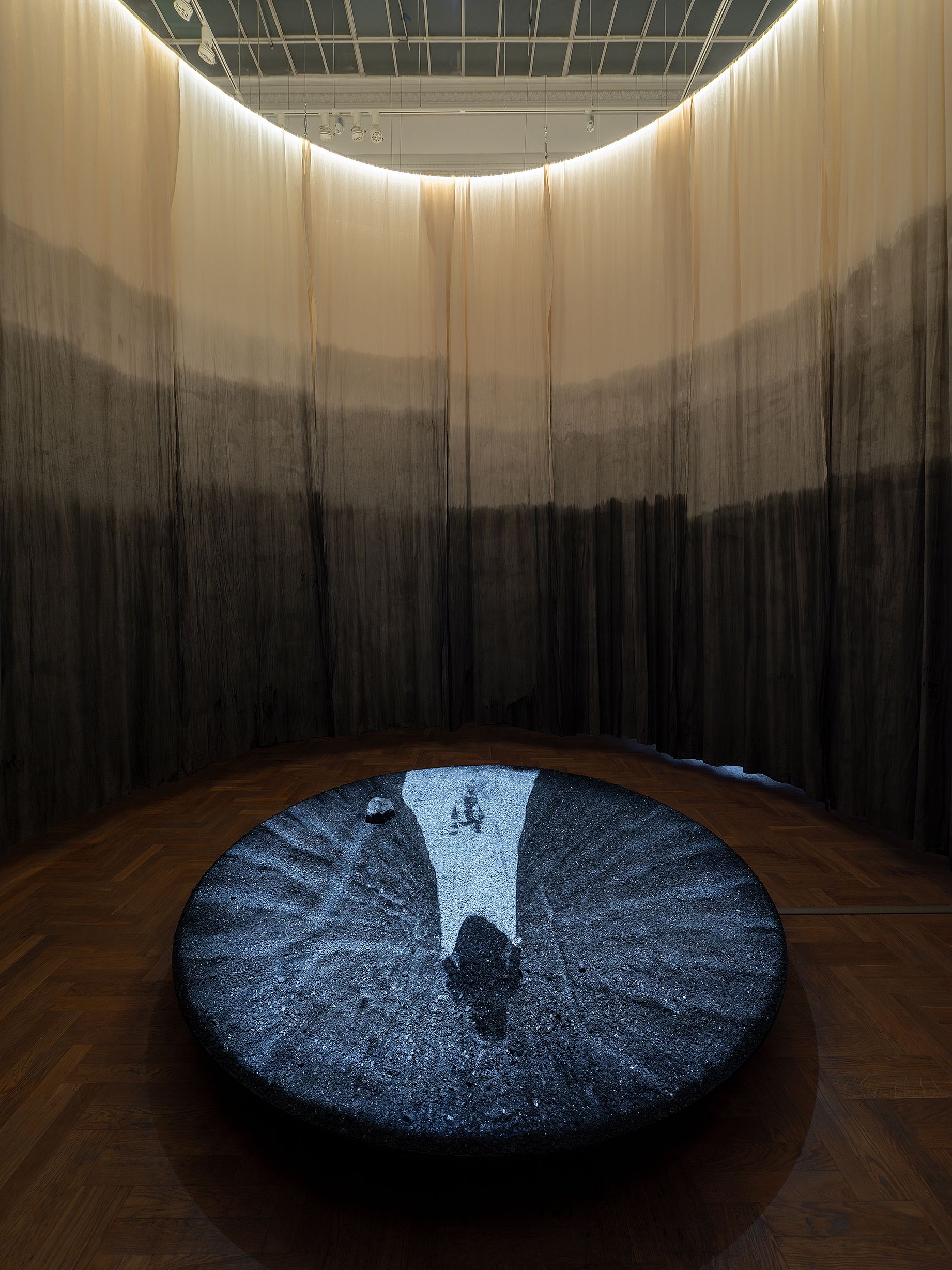 Instalacja artystyczna w galerii przedstawia duży, okrągły, ciemny obiekt na podłodze, otoczony półkolistą, gradientową kurtyną, która przechodzi od jasnego do czarnego.