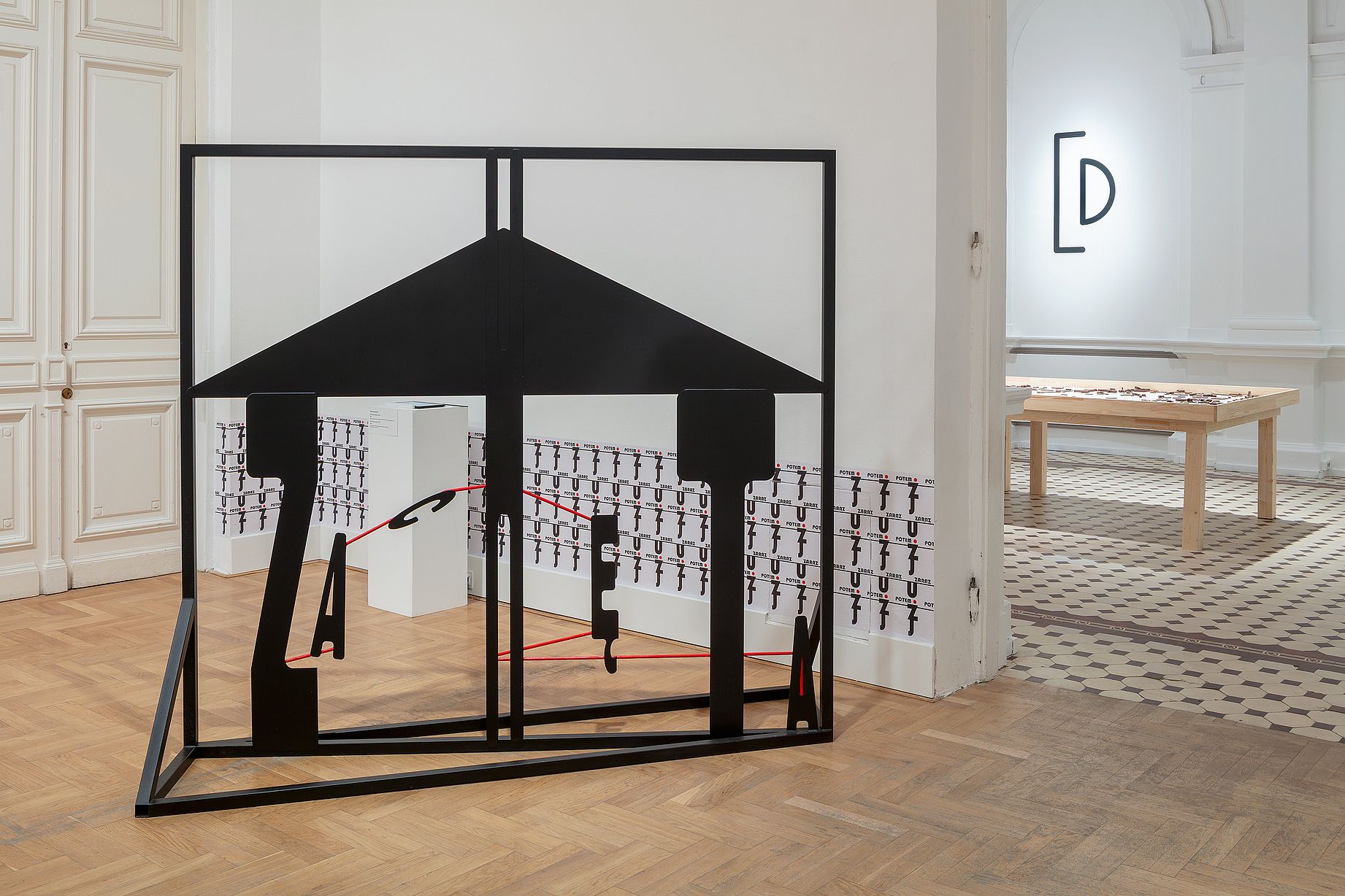 W jasnym wnętrzu galerii stoi duża, czarna, metalowa rzeźba o geometrycznym kształcie przypominającym uproszczoną sylwetkę domu lub bramy tworzącym napis Zachęta, za którą widać białe ściany z drobnymi grafikami oraz przejście do kolejnej sali wystawowej.