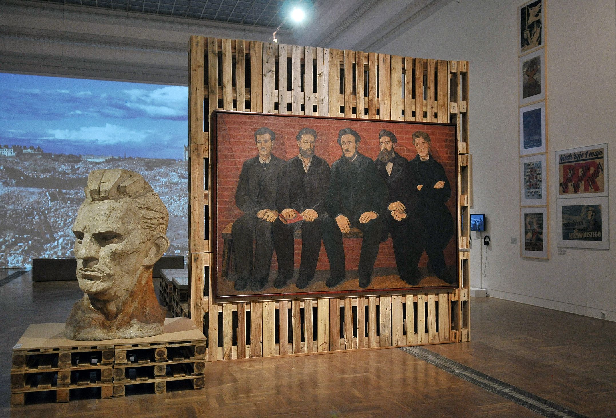Na zdjęciu widać fragment wystawy Zaraz po wojnie w warszawskiej Zachęcie – Narodowej Galerii Sztuki, gdzie obok siebie eksponowane są: po lewej duża, kamienna rzeźba głowy, przypominająca antyczną rzeźbę lub socrealistyczne popiersie, ustawiona na drewnianych paletach, a po prawej stronie tego samego pomieszczenia obraz olejny przedstawiający pięciu mężczyzn siedzących na ławce, również oprawiony w konstrukcję z palet, na tle ściany z propagandowymi plakatami z epok