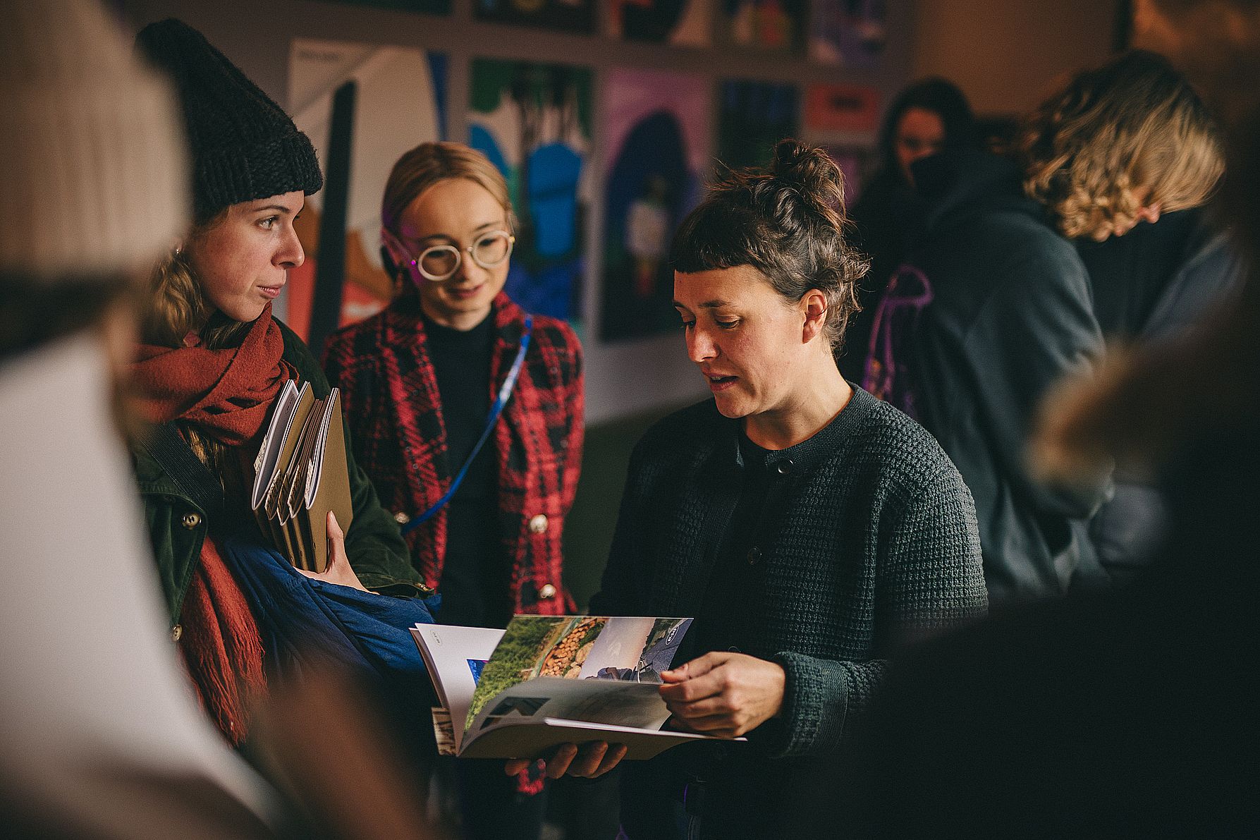 Na zdjęciu kilka osób ogląda wspólnie książkę artystyczną podczas spotkania w galerii, rozmawiając i dzieląc się wrażeniami.
