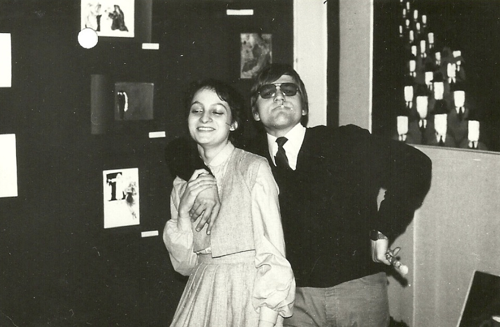 Na czarno-białym zdjęciu młoda kobieta uśmiecha się z zamkniętymi oczami, a obok niej stoi mężczyzna w okularach przeciwsłonecznych i garniturze, obejmując ją ramieniem w żartobliwym geście.