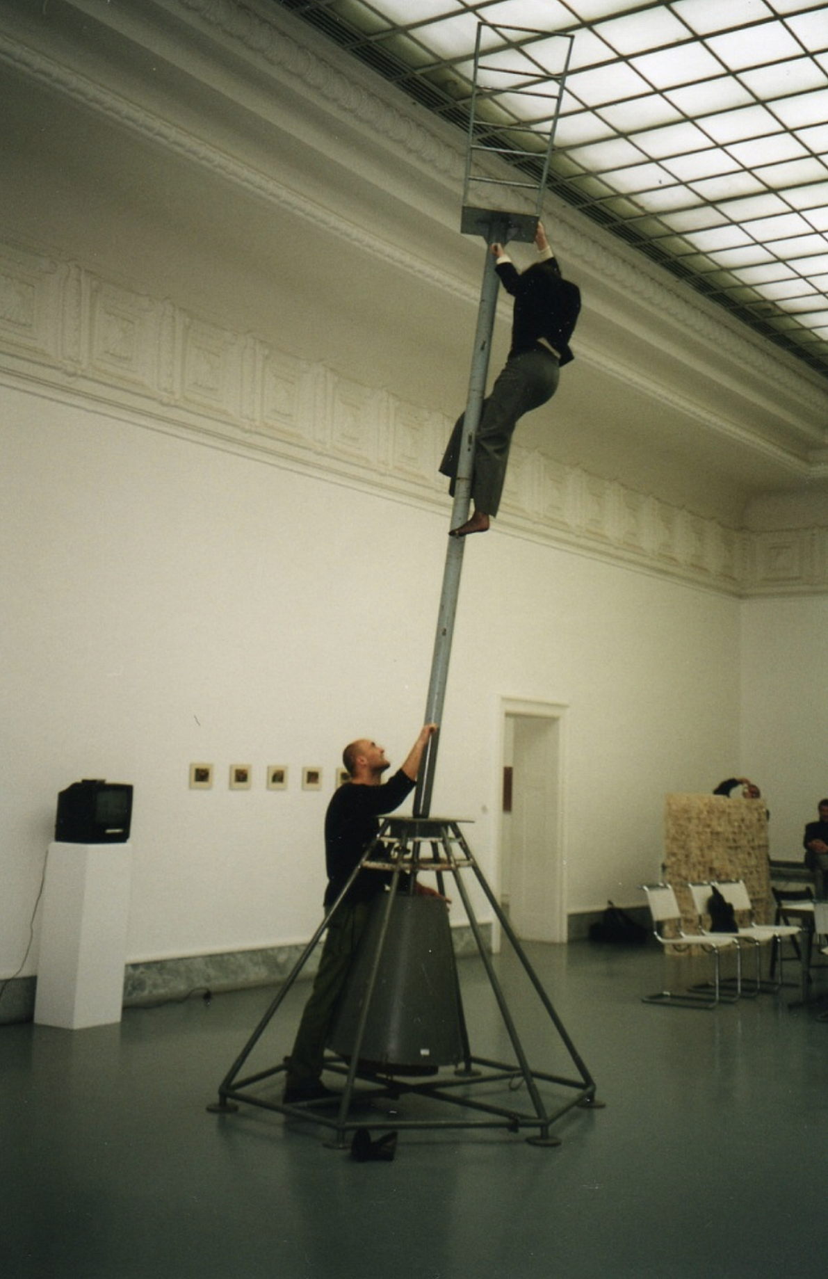 Na zdjęciu widać dwie osoby w galerii sztuki – jedna wspina się wysoko po metalowej konstrukcji przypominającej drabinę, a druga stoi na dole i ją podtrzymuje.