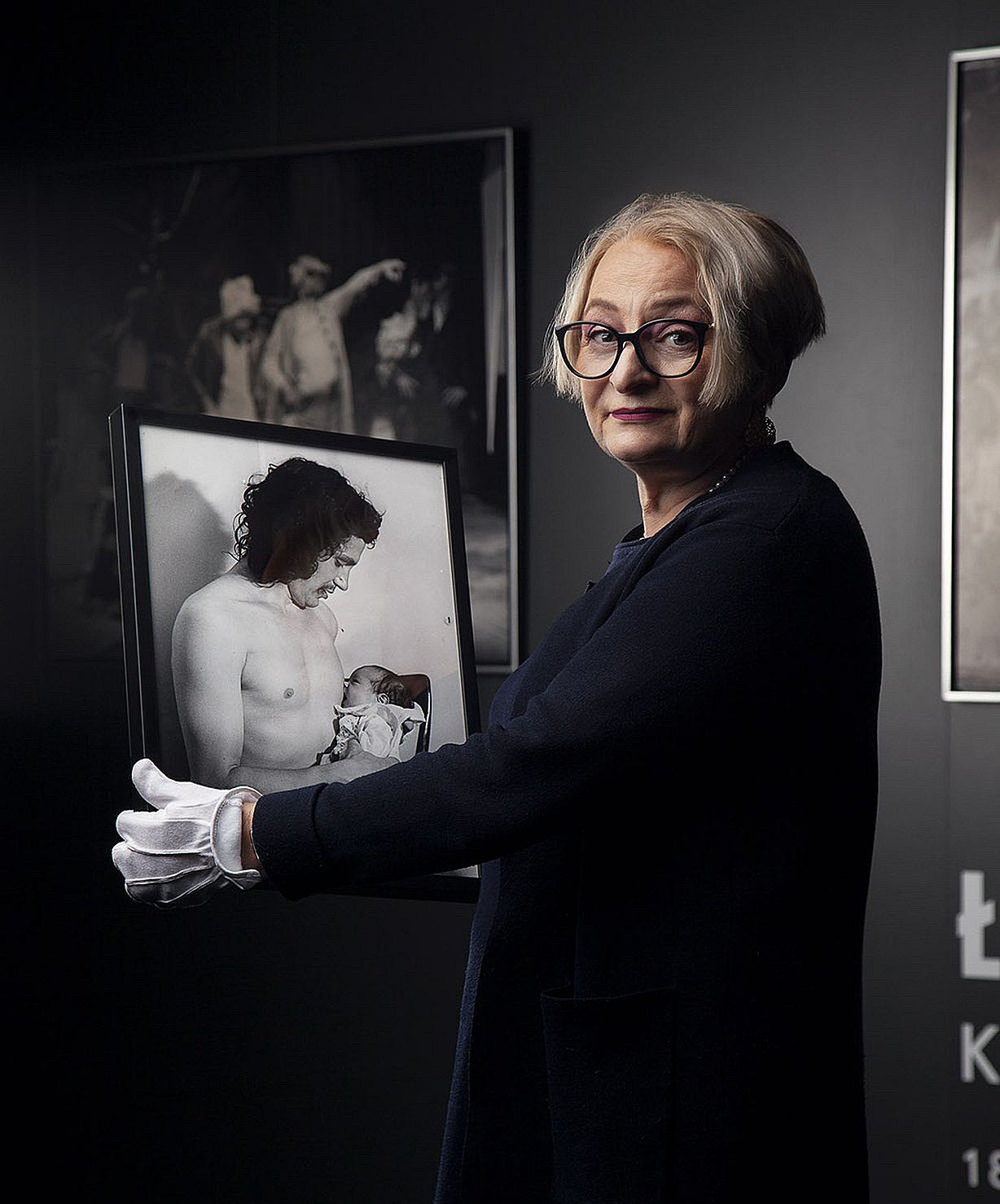 kobieta w okulrach, ubrana na czarno, trzyma w reku fotografię mężczyzny trzymajacego dziecko