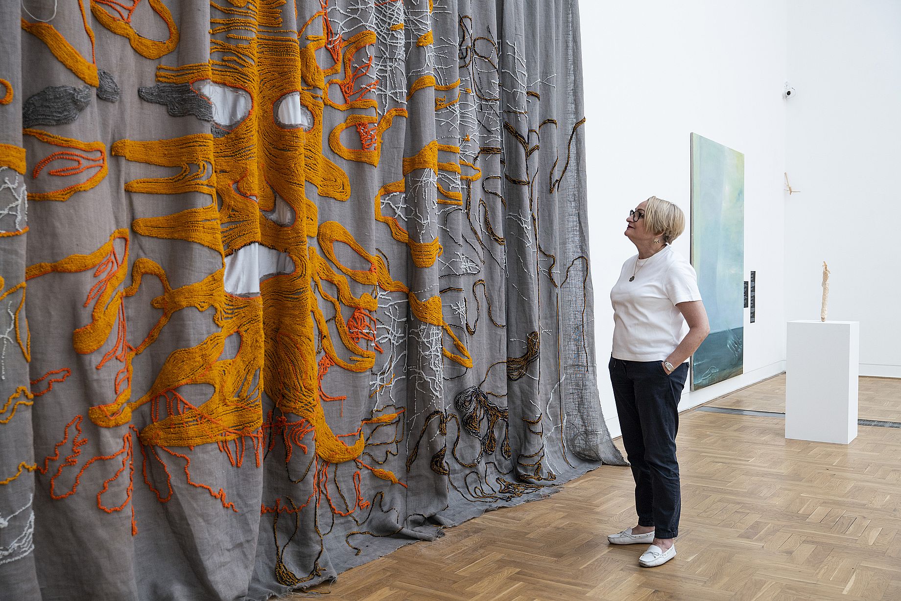 Kobieta w okularach, białych butach stoi przed wielką artystyczną tkaniną z pomarańczowymi akcentami