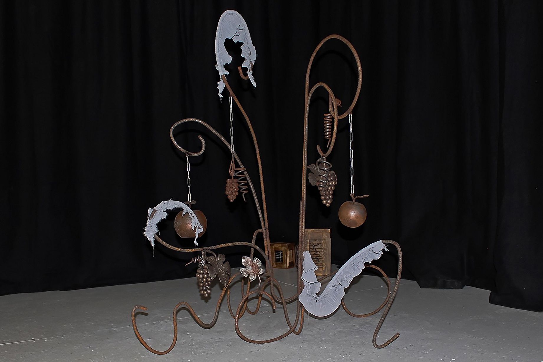 Na zdjęciu przedstawiony jest ozdobny, kuty stojak z metalu i szkła, stylizowany na pnącze winogron, z wiszącymi elementami przypominającymi owoce i liście.