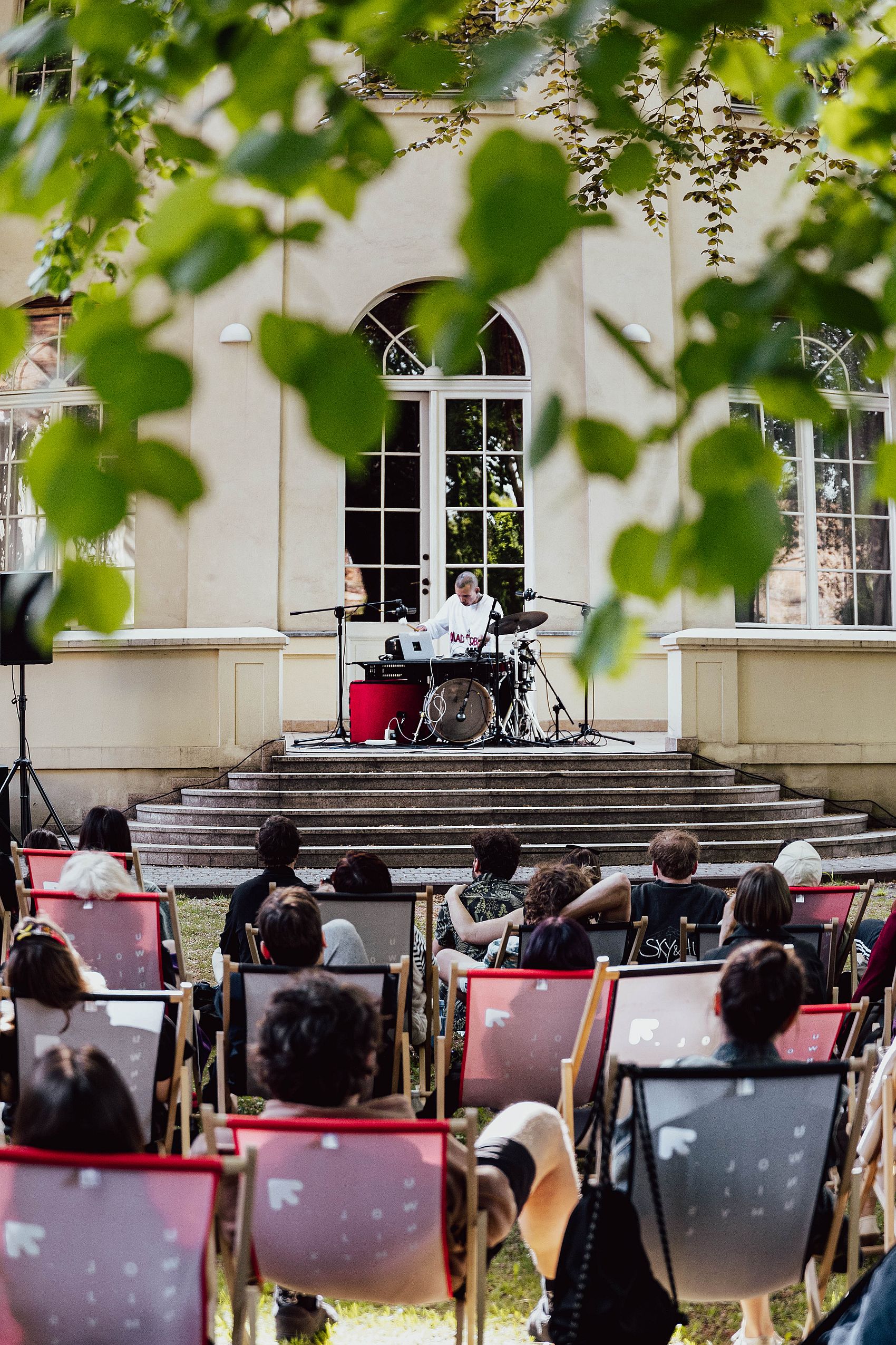 KOncert w pałacowym ogrodzie, publiczność siedzi na leżakach, liście drzewa