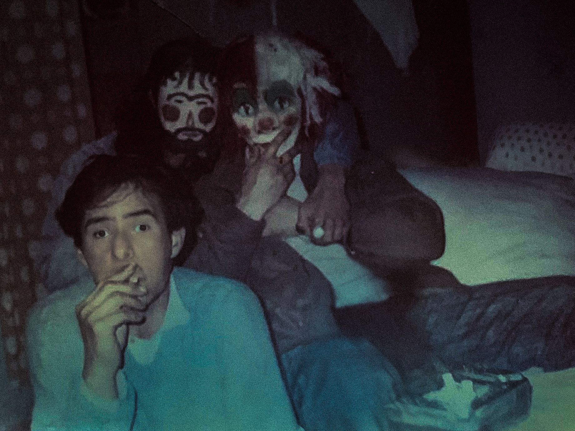 Na słabo oświetlonym zdjęciu widać trzy postacie siedzące na łóżku lub kanapie, z których jedna, w jasnoniebieskiej bluzie, pali papierosa i patrzy wprost na aparat, a dwie pozostałe, za nią, mają na twarzach charakterystyczny, przypominający lalki lub maski makijaż, nadający im nieco upiorny wygląd.