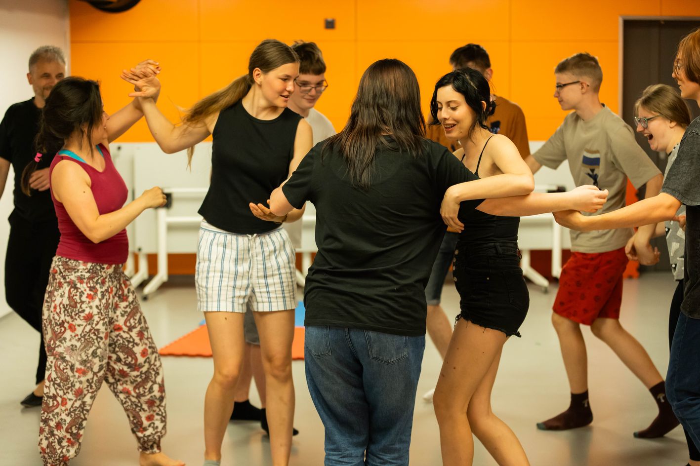 Grupa młodych osób tańczy razem w sali o pomarańczowych ścianach, śmiejąc się i trzymając za ręce w radosnym, integracyjnym kręgu.