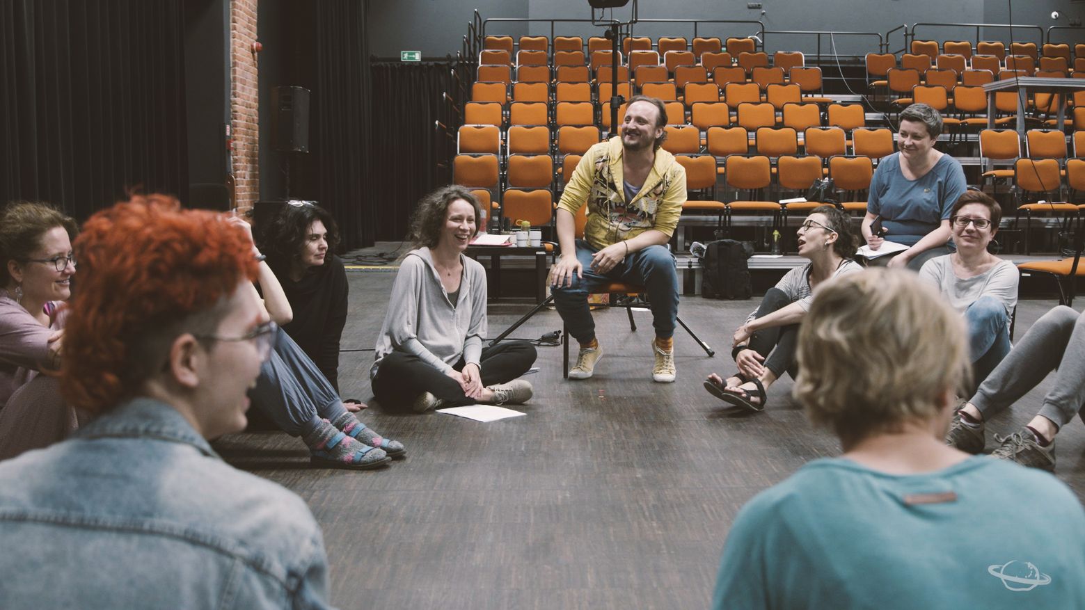 Na zdjęciu widać grupę osób siedzących w kręgu na scenie teatralnej, uśmiechniętych i zaangażowanych w rozmowę; w centrum znajduje się mężczyzna w żółtej bluzie, który najprawdopodobniej prowadzi spotkanie lub warsztat twórczy.