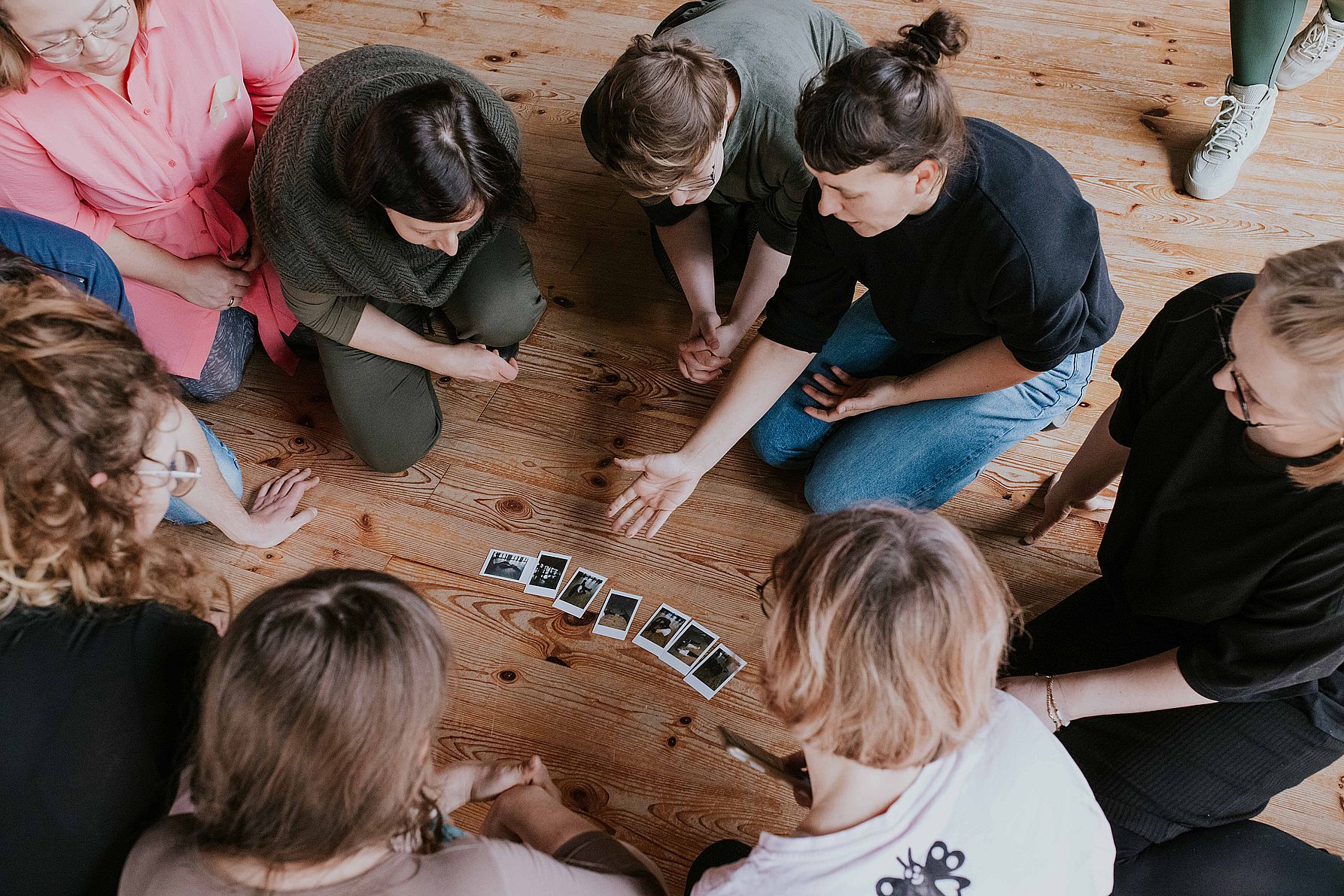 Na zdjęciu kilka osób  siedząc na podłodze ogląda wspólnie zdjęcie podczas spotkania w galerii, rozmawiając i dzieląc się wrażeniami.