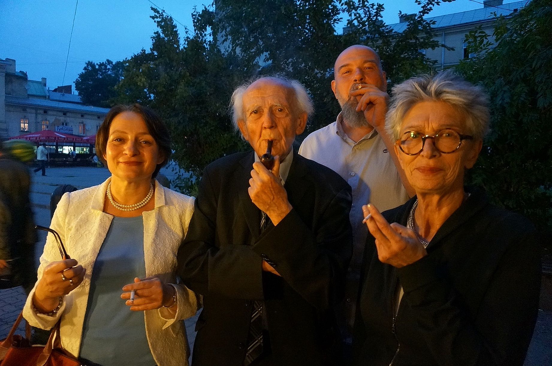 Na zdjęciu wieczorem na zewnątrz stoją cztery uśmiechnięte osoby, z których trzy trzymają fajki lub papierosy.